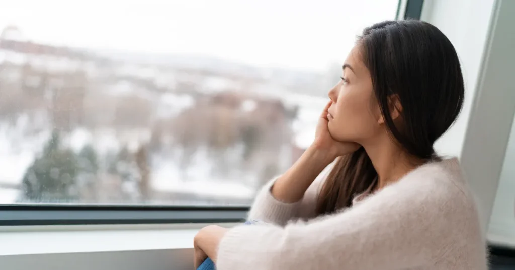 Mulher jovem sentada próxima à janela, olhando com expressão pensativa e introspectiva para uma paisagem coberta de neve no inverno, ilustrando os sentimentos de melancolia e isolamento causados pela depressão sazonal.